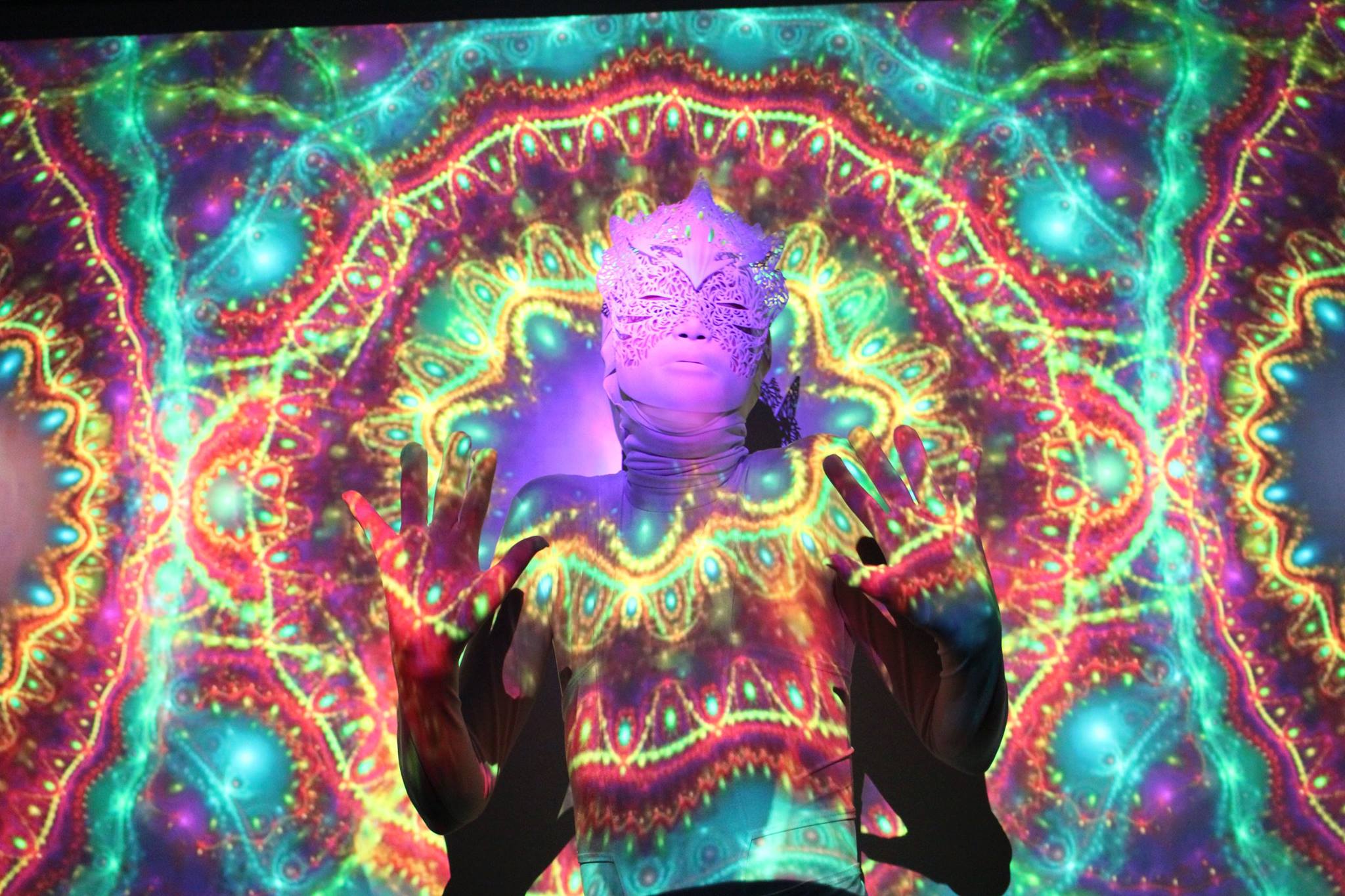 A person wearing an ornate mask standing with hands up as a bright and colourful design is projected onto them.