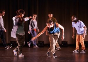 A group people dressed in sportswear performing on stage. One walks with outstretched arms.