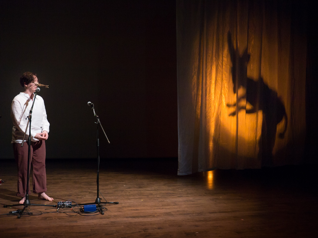 A person performing on stage. The their left in shadow on the curtain appears to be a monster.