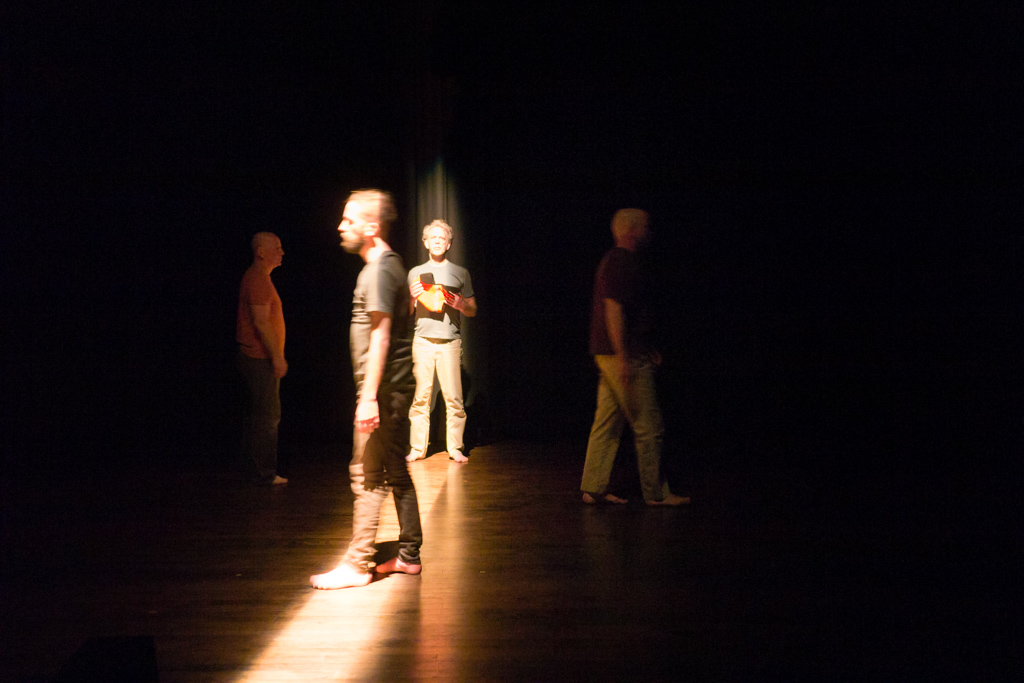 4 performing on a dimly lit stage. A shaft of light casts down the middle.