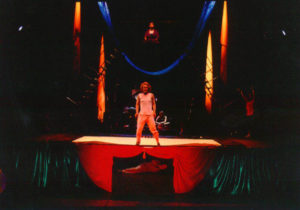 A man stands on stage with a defiant stance. Beneath the stage someone lies on their side, hiding. Above the stage sits a man in a robe, dramatically lit.