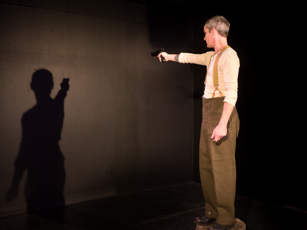 Performer Ryan Kerr, dressed in military slacks, stands on a stool aiming a gun at his shadow on the black wall