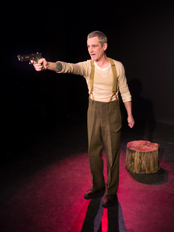 Performer Ryan Kerr stands in a black room lit with a red spotlight. He stands in front of a stump, aiming a handgun forward
