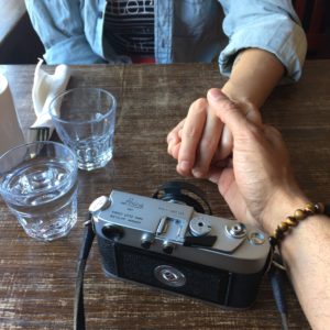photo of two people at a table on holding the other's hand. a camera on drinking glasses and cutlery are on the table.