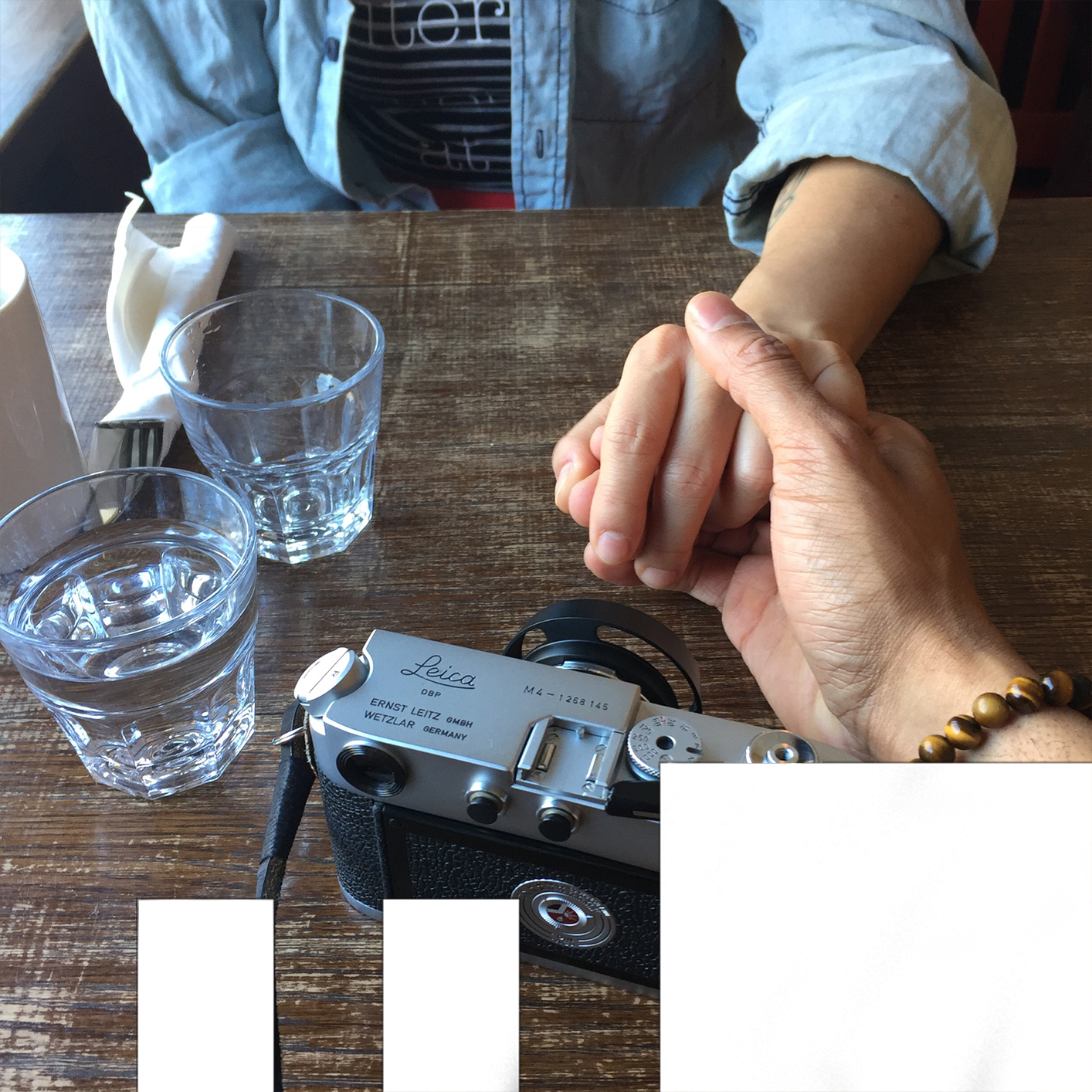 photo of two people at a table on holding the other's hand. a camera on drinking glasses and cutlery are on the table.