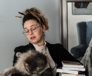 A woman with pens in her hair, books on her lap and a Cat trying to escape up from her arms.