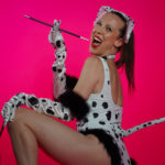Individual wearing Dalmatian bodysuit, ears, gloves, and a leash in crouching position looks at camera with big smile, holding long cigarette holder. Pink backdrop.