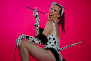 Individual wearing Dalmatian bodysuit, ears, gloves, and a leash in crouching position looks at camera with big smile, holding long cigarette holder. Pink backdrop.