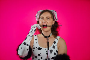 Individual wearing Dalmatian bodysuit, ears, gloves, and a leash is biting long cigarette holder like a dog. Pink backdrop.