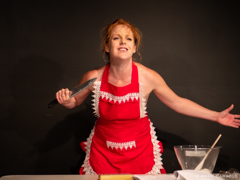 Individual wearing red apron with white lace holds knife in one hand with other arm outstretched, mixing bowl and spoon on table in front.