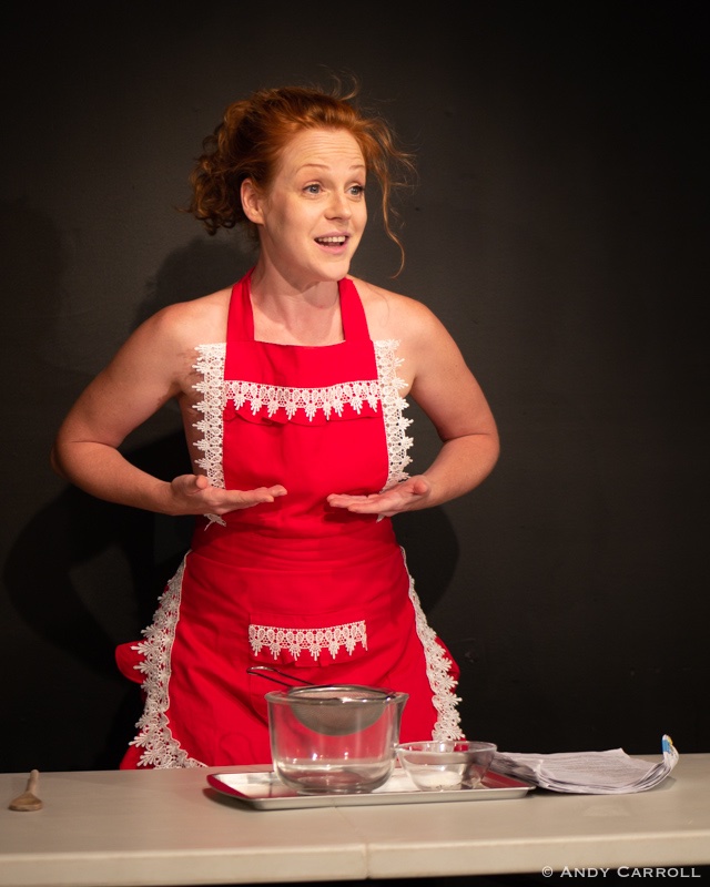 Individual wearing red apron with white place palms of hands under chest, as if holding breasts, eyebrows raised, almost smiling. Spoon, sifter, mixing bowls, and cookie sheet on table in front.