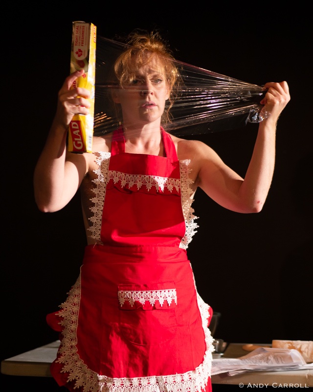 Individual wearing red apron with white lace holds saran wrap in front of their face, one hand holding the box and the other pulling the saran wrap taut. Individual looks at saran wrap with empty expression.