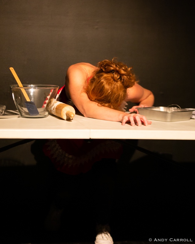 Individual sprawls out on table with baking supplies, one arm outstretched on the table, face down. A mixing bowl, spatula, rolling pin and baking dish surround them.