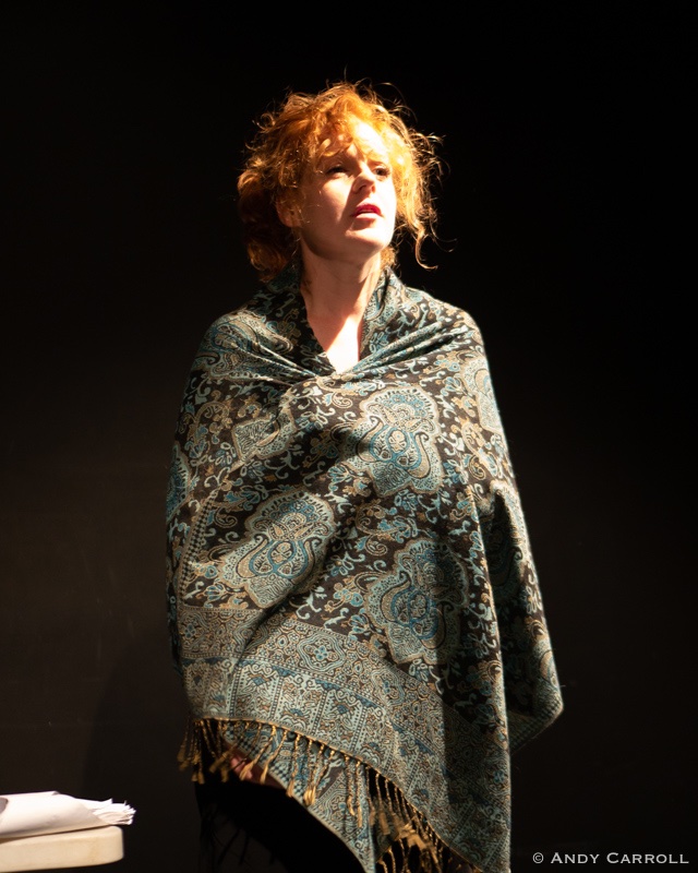 Individual with disheveled red hair wears patterned blue and grey blanket wrapped around shoulders and looks off into the distance.