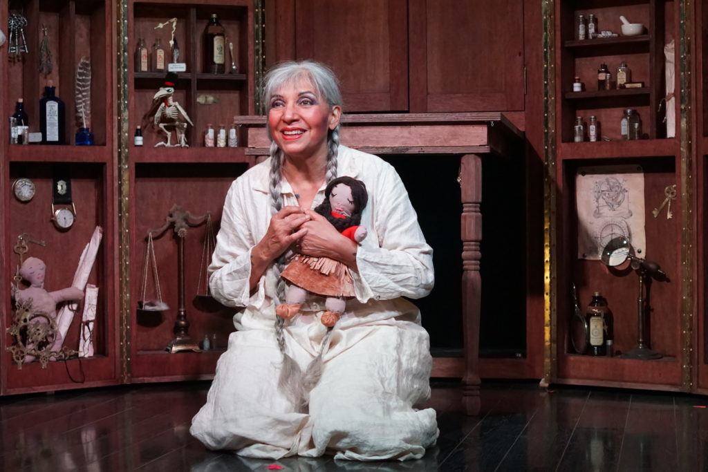 Performer Monique Mojica crouches in a white nightgown clutching a doll.