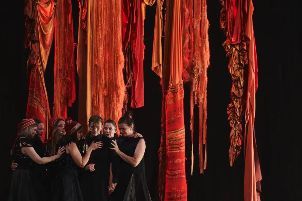 Seven women, hands on eachothers shoulders, perform in front of a vibrant red textile backdrop