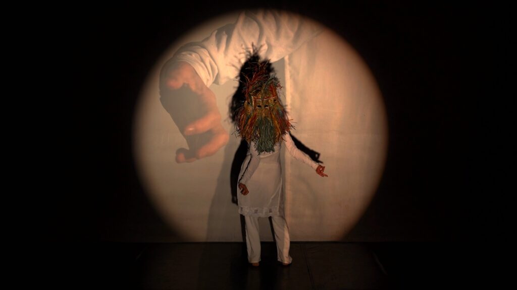 Norma Araiza performs in front of a projection of a hand and arm. She faces away from the audience, but wears a mask facing towards them, making it appear that her head or body is on backwards.