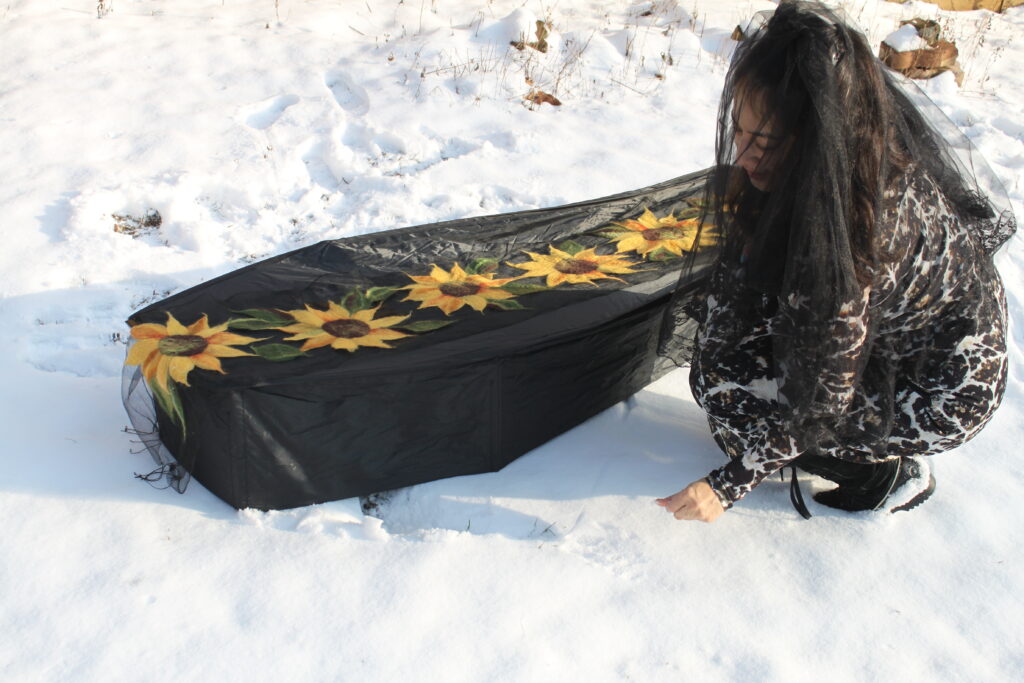 Heryka Miranda crouches by a coffin draped in textile sunflowers. She wears a black veil. There is snow on the ground.