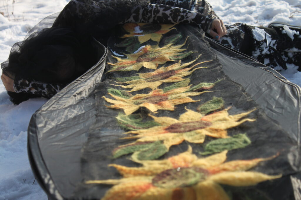 Heryka Miranda is curled in the fetal position at the head of a coffin draped in textile sunflowers. She wears a black veil. There is snow on the ground.