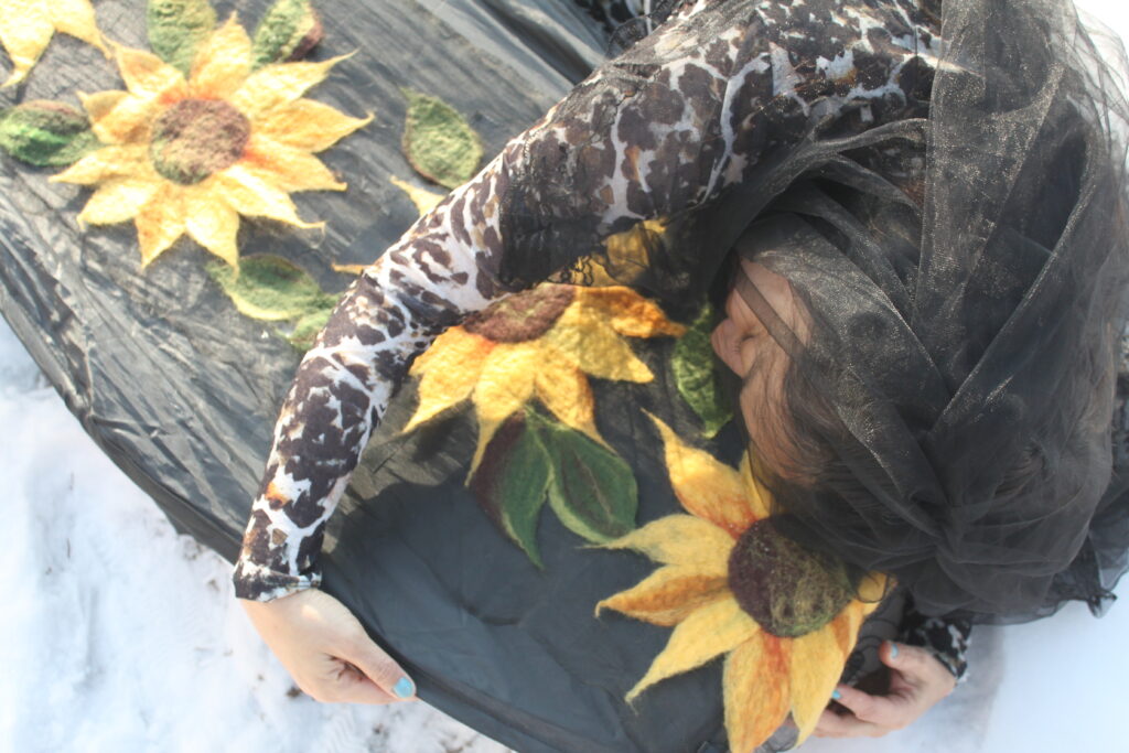 Heryka lays in the snow hugging a coffin draped in textile sunflowers