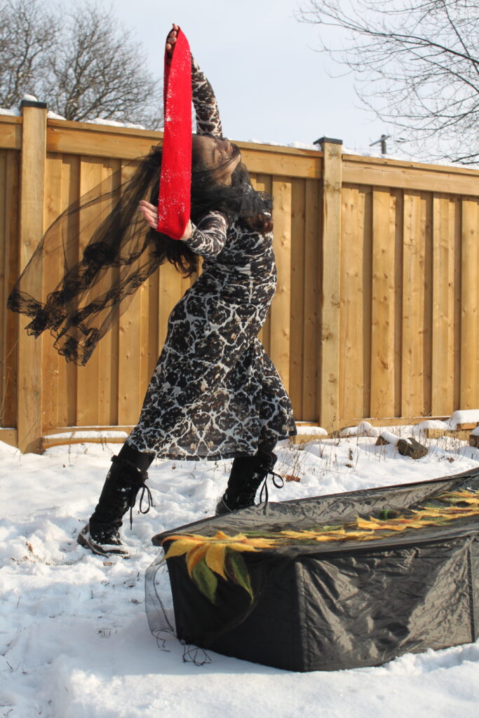 Heryka dances in the snow beside the coffin. She wears a brown black and white printe dress and a black veil. She stretches a red strip of fabric between her hands.