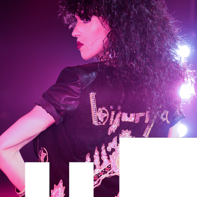 Bijuriya Gabriel Dharmoo performs as Bijuriya, wearing a voluminous curly wig and a short sleeved leather top with Bijuriya written across the back. The image is bathed in pink light. in the photo.