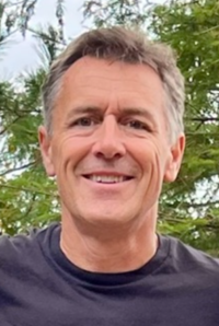 Headshot of a man with short greyish brown hair smiling. There are trees in the background.