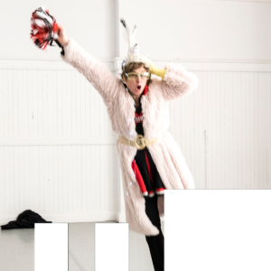 Sara Porter stands with her hand pointing upwards, she wears.a furry pink robe, feathers in her hair, yellow glasses and a silly expression of surprise on her face
